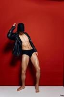 nude man in shorts and leather jackets posing on a red background and a black mask on his head photo