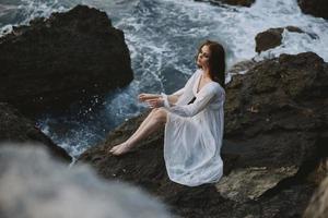 bonito mujer con mojado pelo en un blanco vestir en el costa se sienta en el rocas foto