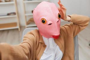 Funny blonde lady in pink fish mask warm sweater doing selfie use phone sitting in armchair at modern home interior. Pause from work, take a break, social media in free time concept. Wide angle photo