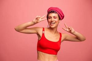 joven atlético mujer con un corto Corte de pelo y púrpura pelo en un rojo parte superior y un rosado sombrero con un atlético figura sonrisas y muecas mirando a el cámara en un rosado antecedentes foto