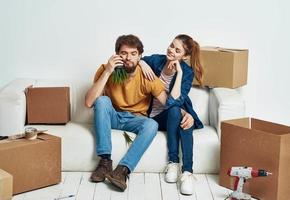 man and woman sitting on a white sofa in a room with boxes of things tools moving photo