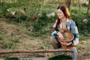 un mujer granjero sonrisas y vierte comida para el aves dentro un pájaro alimentador desde un cuenco a un al aire libre granja a aumento saludable, orgánico pollos foto