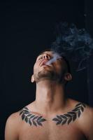 Modelling snapshots. Pensive focused tanned attractive handsome naked man tipping his head back smoking posing isolated in black studio background. Fashion offer. Copy space for ad. Closeup photo