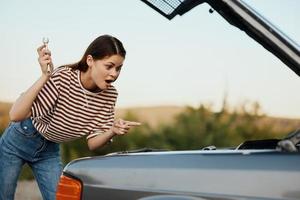 A woman with a wrench with a smile looks happily under the open hood of her car and repairs it from a roadside breakdown while traveling alone in nature photo