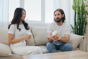 hombre y mujer sentado a hogar en el sofá en blanco elegante camisetas Bebiendo café fuera de cangrejo tazas desde un café tienda y teniendo divertido chateando sonrisas y la risa a hogar. masculino y hembra amistad foto