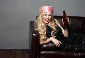 A beautiful blonde with a bottle of beer lies on a leather sofa indoors and a pink mask for sleeping photo