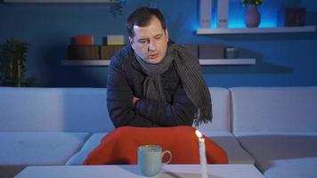 homem sozinho dentro frio casa bebendo quente bebida, sentado de luz de velas. homem sentado em sofá dentro sem aquecimento frio casa dentro inverno com casaco e lenço. video