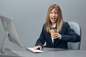 Overjoyed millennial blonde businesswoman worker in blue jacket using desktop computer with takeaway coffee in gray modern office. Remote Job, Technology And Career Profession Concept. Copy space photo