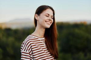 de cerca retrato de un joven mujer con un hermosa sonrisa con dientes en un a rayas camiseta en contra el antecedentes de arboles foto