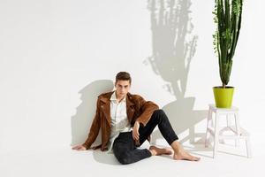 handsome man in brown jacket and trousers sits on the floor near a flower in a pot photo