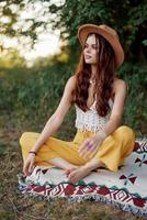 mujer en ecológico ropa en un hippie Mira se sienta en un de colores tartán meditando en el loto posición sonriente y mirando a el otoño puesta de sol en el parque foto