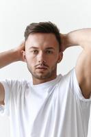 Closeup portrait shot of handsome tanned man guy in basic t-shirt holds hands behind head looks at camera posing on white background. Fashion Style New Collection Offer. Copy space for ad. Model snap photo