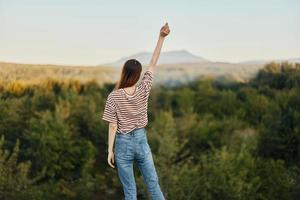 un joven mujer soportes con su espalda a el cámara con su manos arriba en un camiseta y pantalones en naturaleza y disfruta un hermosa ver de el montañas. otoño viaje a naturaleza estilo de vida foto
