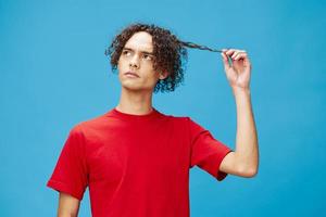 Pensive thoughtful unhappy young curly tanned Caucasian guy in basic red t-shirt posing isolated on over blue background. Lifestyle and Emotions concept. Good offer with copy space for ad photo