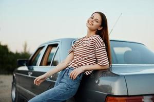 un joven mujer en un a rayas camiseta y pantalones soportes por su coche descansando desde conduciendo, propensión en su coche junto a el la carretera y sonriente foto