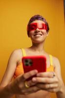 Woman blogger smiling in unusual red millennial glasses holding her phone and looking at the screen against an orange background in a stylish yellow top photo