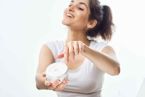 cheerful brunette in a white t-shirt with a jar of cream in her hands skin care light background photo