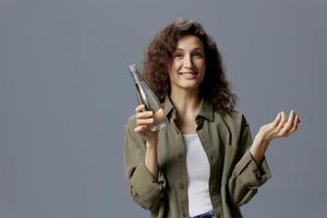 Joyful smiling curly beautiful woman in casual khaki green shirt hold bottle drink clean water posing isolated on over gray blue background. Healthy lifestyle. Water is Life concept. Copy space photo