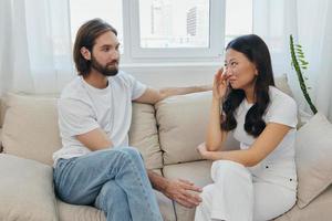 A man and a woman of different races sit on the couch in a room at home and talk about their problems to each other. A stress-free lifestyle of family quarrels with psychological support photo