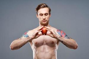 a guy with a pumped-up torso and a tattoo holds an apple in his hands on a gray background photo