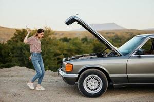 A woman opens the hood of a broken down car and tries to find the cause of the breakdown on a road trip alone and kicks the car in anger photo