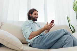 un hombre con un barba se sienta en el sofá durante el día y sonrisas a hogar y mira a su teléfono relajante en su día apagado, vida en línea en su teléfono mensajes de texto con amigos foto