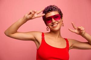 Fashion portrait of a woman with a short haircut in colored sunglasses with unusual accessories with earrings smiles on a pink bright background shows her hands to the camera photo