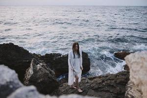 A woman in a white dress stands on rocky stones by the ocean photo