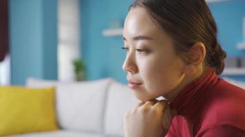 Thoughtful and unhappy Asian woman sitting in living room. Young Asian woman sitting alone at home looking around thoughtfully. video