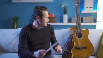 Male musician sitting on the sofa at home playing the violin and composing a new song, playing a song he knows. video