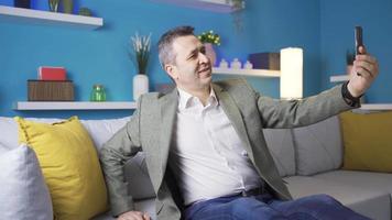 Conceited mature man taking selfie on smartphone alone at home. Man in suit sitting on sofa at home taking pictures on his smartphone, smiling, posing. video