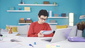 masculino aluna estudo, fazendo trabalho de casa, usando computador portátil, pensamento, questão responder. masculino aluna estudando às dele escrivaninha às lar, levando notas, olhando às computador portátil, pensando sobre ideia. video