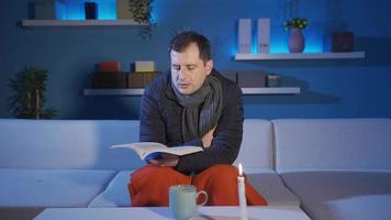 The man sitting on the sofa at home is reading a book by candlelight, he is cold in the cold, there is no gas, the house does not heat up. video
