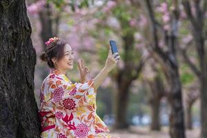 Japanese woman in traditional kimono taking selfie with peace gesture while walking in the park at cherry blossom tree during spring sakura festival photo