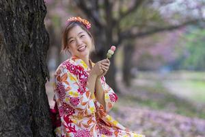 japonés mujer en tradicional kimono vestir participación el dulce hanami dango postre mientras caminando en el parque a Cereza florecer árbol durante primavera sakura festival con Copiar espacio foto