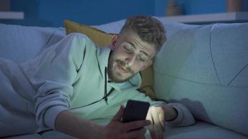 Handsome young man using phone at home in a sad and depressed mood. Unhappy and lonely young man looking at his phone in a helpless and reluctant mood at home, feeling boring, depressed. video