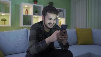 contento joven hombre utilizando teléfono a hogar, agradable y divertido. joven hombre riendo a el social medios de comunicación mensajes y publicaciones él gustos en el teléfono. video