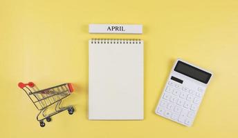 flat layout of opened blank page notebook with wooden calendar April, shopping trolley or shopping cart and white calculator on yellow background. monthly financial plan concept. photo