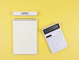 plano diseño de abrió blanco página cuaderno con de madera calendario abril y blanco calculadora en amarillo antecedentes. mensual financiero plan concepto. foto