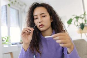 Asian woman using cotton swab while doing coronavirus PCR test. Woman takes coronavirus sample from her nose at home. woman at home using a nasal swab for COVID-19. photo