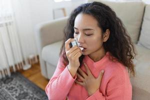 asiático mujer utilizando inhalador mientras sufrimiento desde asma a hogar. joven mujer utilizando asma inhalador. de cerca de un joven asiático mujer utilizando asma inhalador a hogar. foto