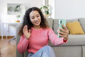 atractivo sonriente asiático mujer utilizando inteligente teléfono mientras sentado en el sofá a hogar. comunicación y vídeo llamada concepto. foto