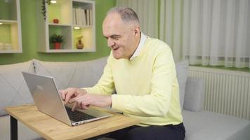Happy old man is happy to do his work on laptop at home in an easy and practical way. The old man who does his online transactions without leaving home is happy and cute. video