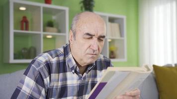 äldre man med öga oordning bär glasögon medan läsning en bok. gammal man med visuell nedsättning har syn problem och bär glasögon medan läsning en bok på Hem. video