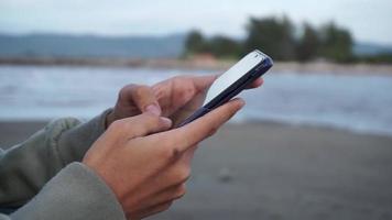 mano participación teléfono inteligente y mecanografía. hombre mano chateando con teléfono de mano. utilizando teléfono inteligente cerca el playa video