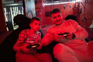 Father and son play gamepad video game console in red gaming room. Dad and kid gamers. photo