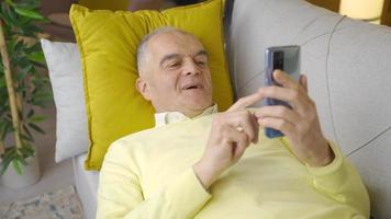 contento vecchio uomo dire bugie su divano utilizzando Telefono. rilassato spensierato pensionato vecchio uomo dire bugie su divano a casa guardare a Telefono. video