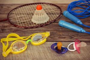 Pacifier and sports equipment the birdie is on the racket, skipping rope, swimming goggles on wooden background. Concept of sports to be engaged with early childhood. Toned image. photo