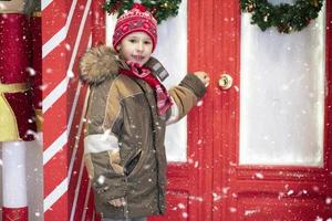 A beautiful child in winter clothes is standing near the red doors. photo