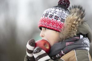 niño en el invierno con fruta. chico en invierno ropa come un rojo manzana foto